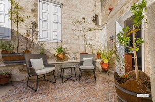 Un patio con muros de piedra, dos sillas de mimbre con cojines, una pequeña mesa y plantas en macetas.