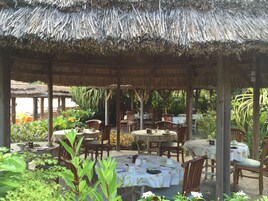 An outdoor dining area with thatched roofs and wooden tables, surrounded by lush greenery.