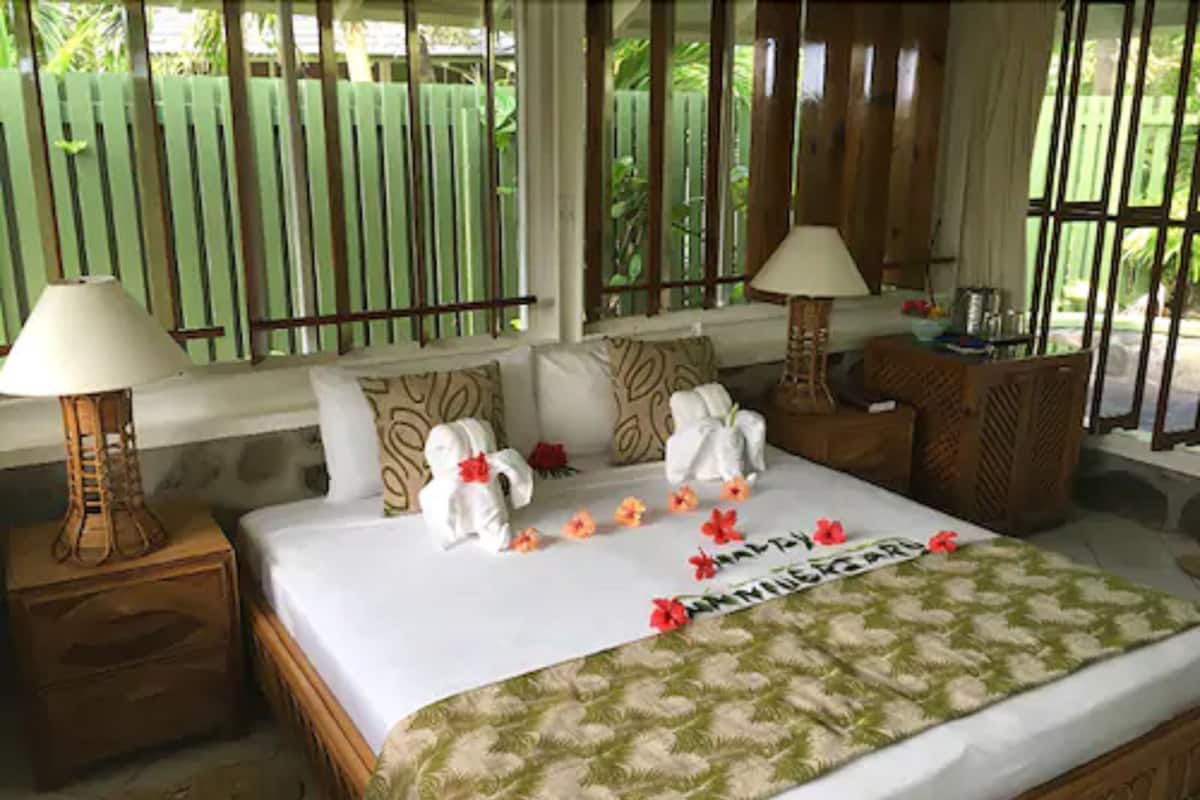 A neatly made bed with towels and flowers, a wooden headboard, and a window with a view of greenery.
