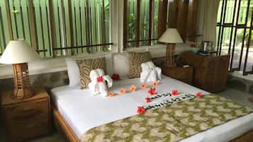 A neatly made bed with towels and flowers, a wooden headboard, and a window with a view of greenery.