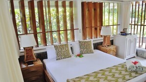 A hotel room with a large bed, wooden headboard, two bedside tables with lamps, a small bedside table with a lamp, a window with vertical blinds, and a view of greenery outside. - Young Island Resort (Villa)