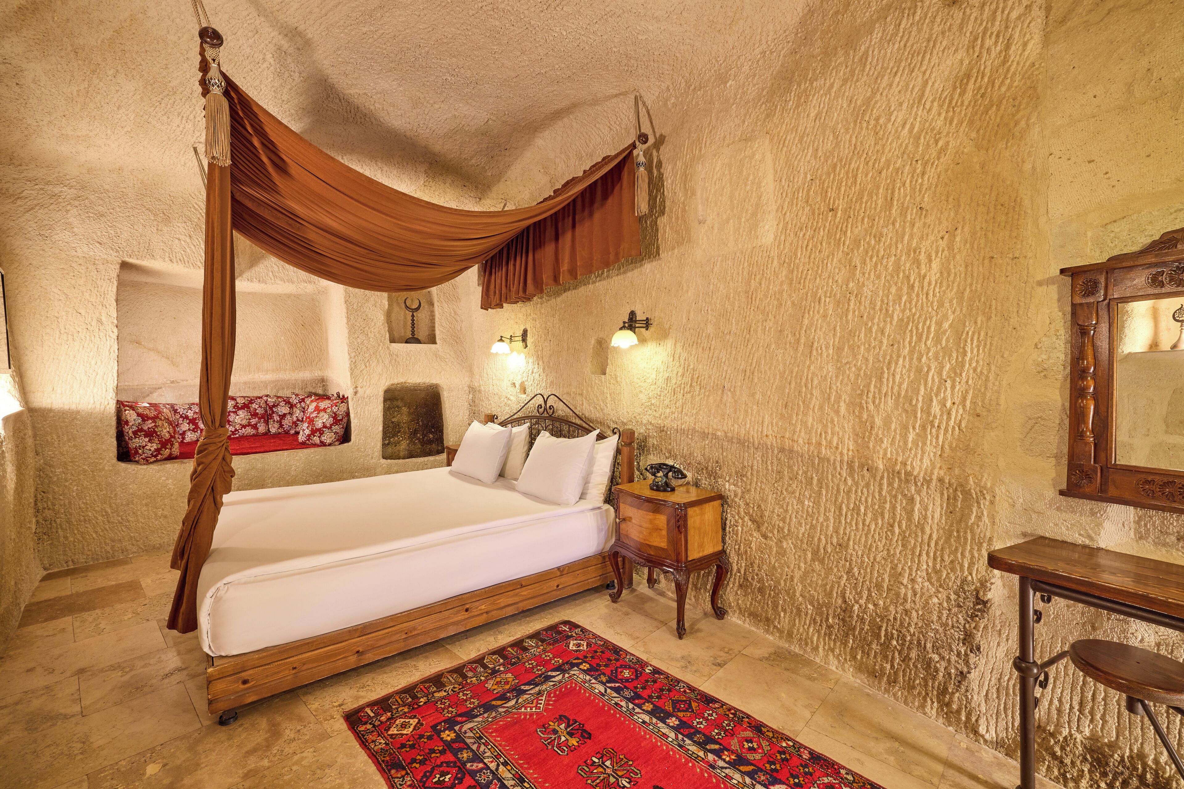 A bedroom with a four-poster bed, a nightstand, a wooden desk, and a red patterned rug.