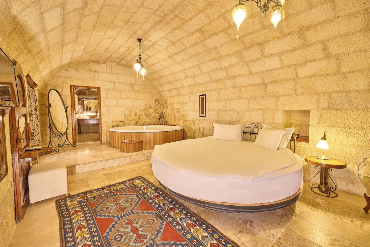 A bedroom with a large bed, a wooden bench, a round bathtub, and a small table with a lamp.