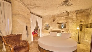 A round bed in a stone-walled room with a vintage armchair and a small table with a decorative item. - Mithra Cave Cappadocia (Nevsehir)