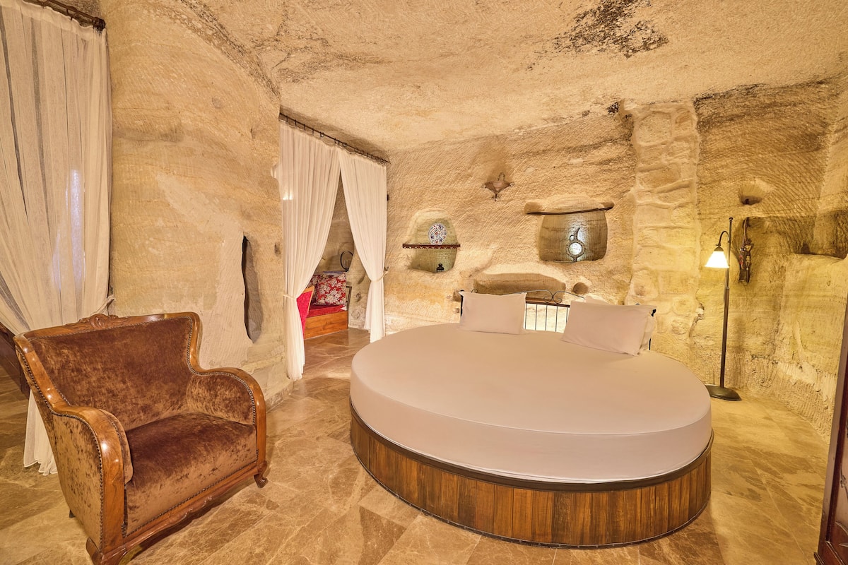 A round bed in a stone-walled room with a vintage armchair and a small table with a decorative item.