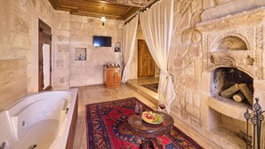 Una habitación de paredes de piedra con chimenea de piedra, una bañera, una mesa de comedor con comida y vino, y un armario de madera.