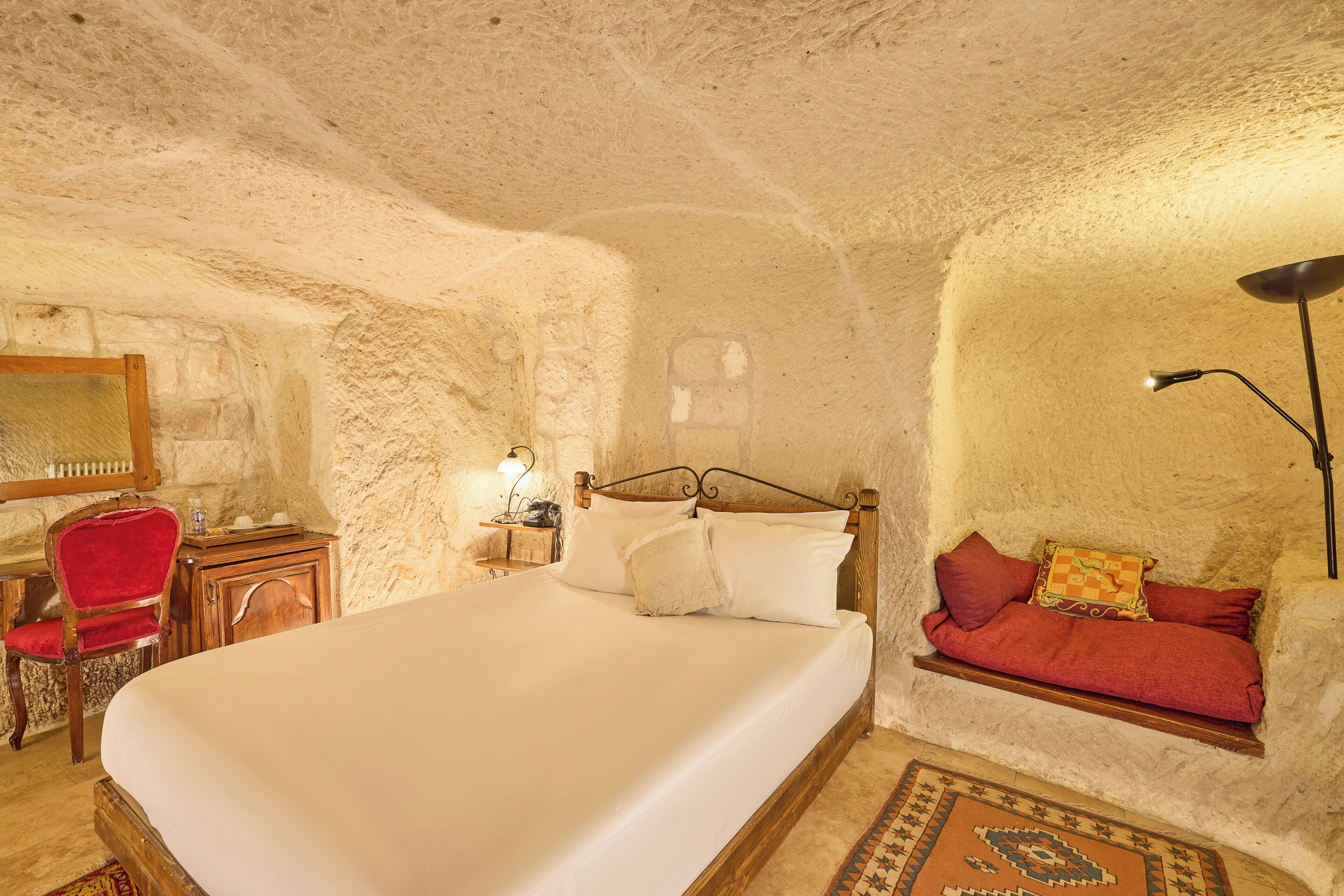 Une chambre avec un grand lit, une table de chevet en bois, une chaise rouge, une petite table avec une lampe et une étagère fixée au mur.