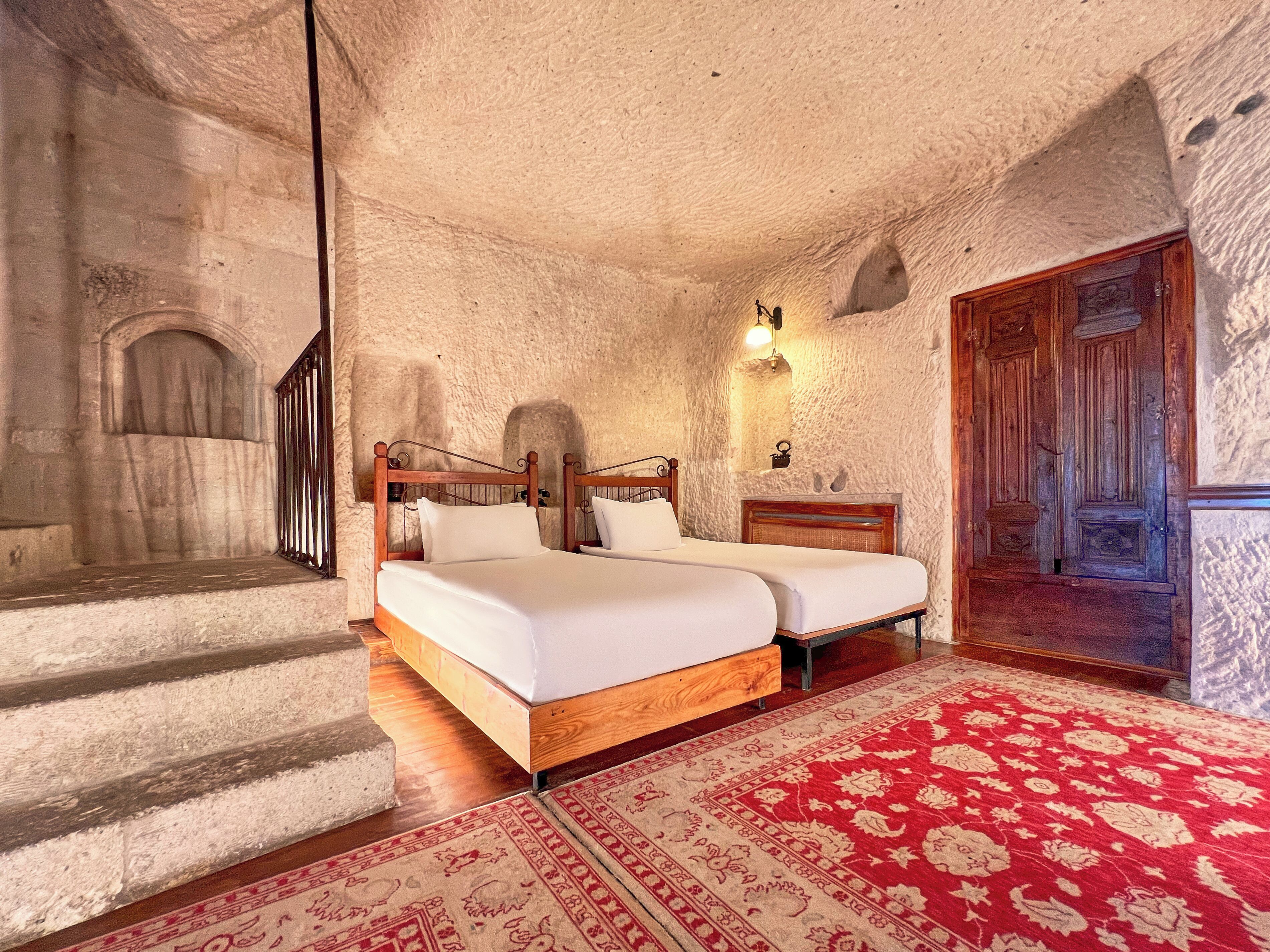 A bedroom with a bed, a nightstand, a wall-mounted light, a wooden door, and a red patterned rug.