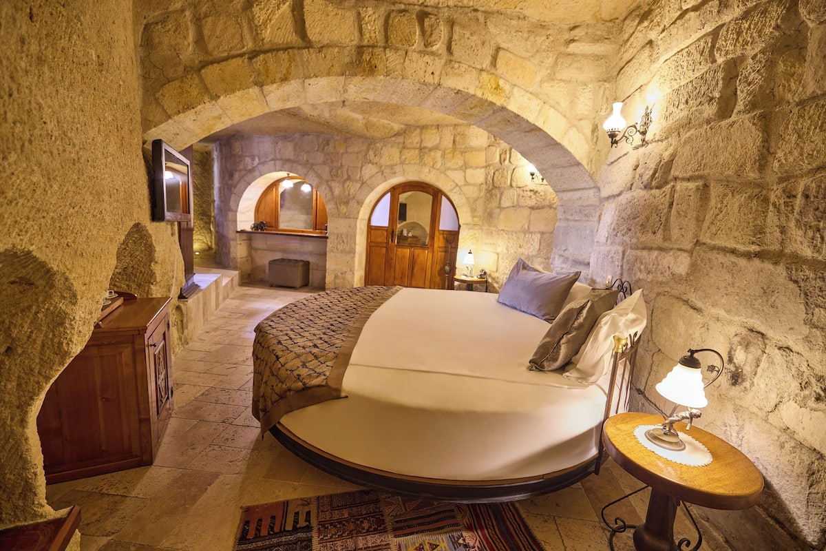 A stone-walled bedroom with a round bed, a wooden dresser, and a small table with a lamp.