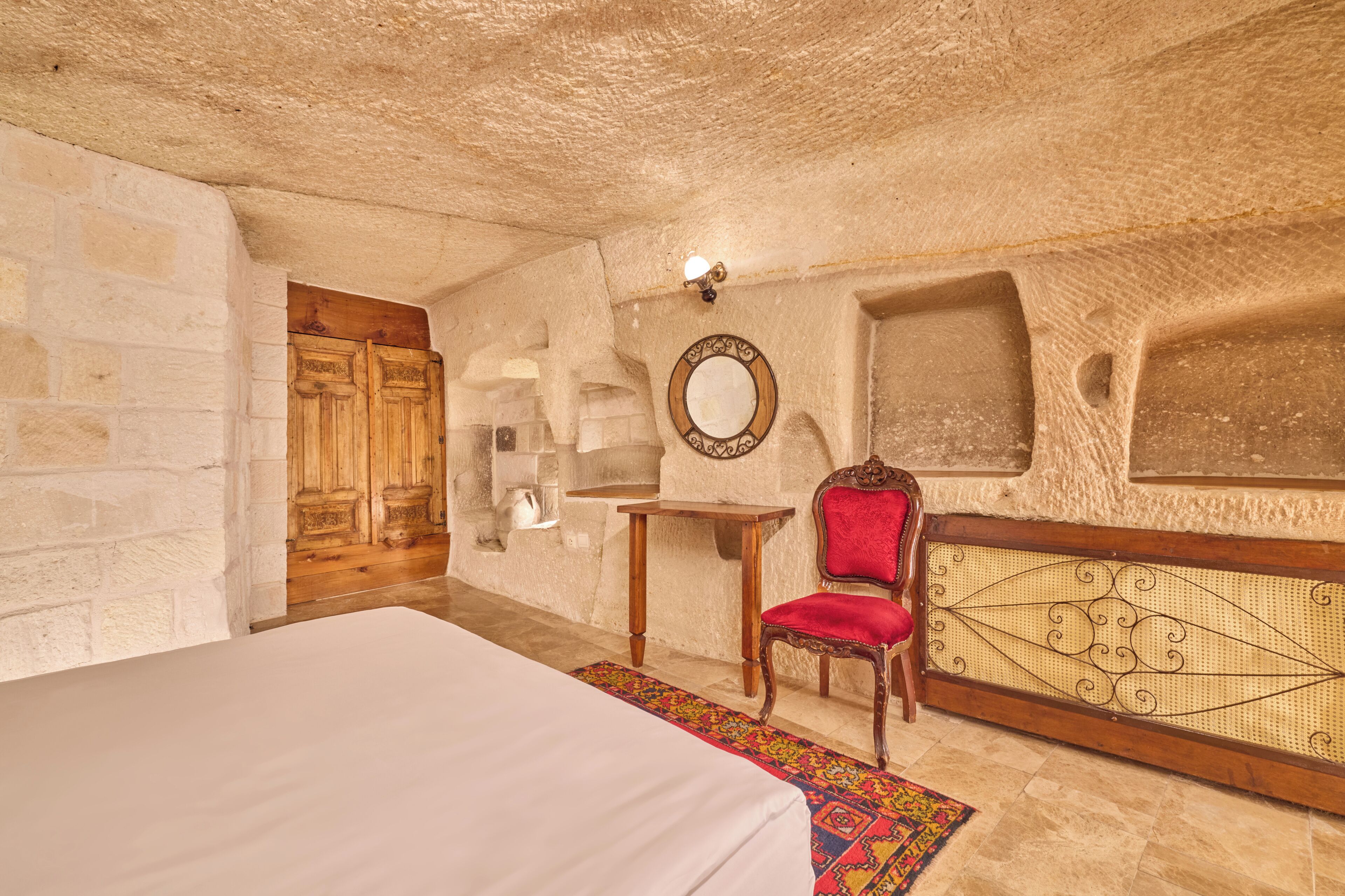 A room with a bed, a wooden chair, a small table, a mirror, and a decorative wall panel.