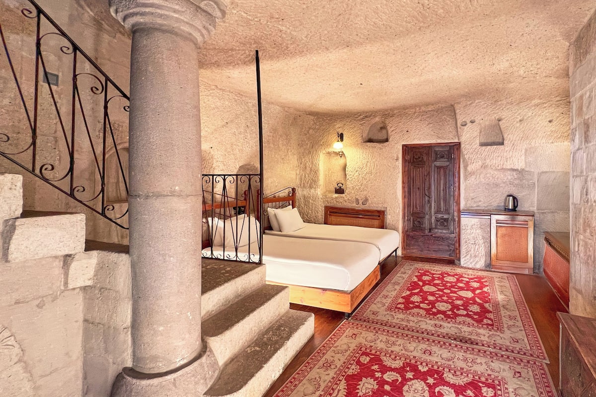 A stone-walled bedroom with a bed, a nightstand, a wooden bench, a red patterned rug, a staircase with a metal railing, and a door.