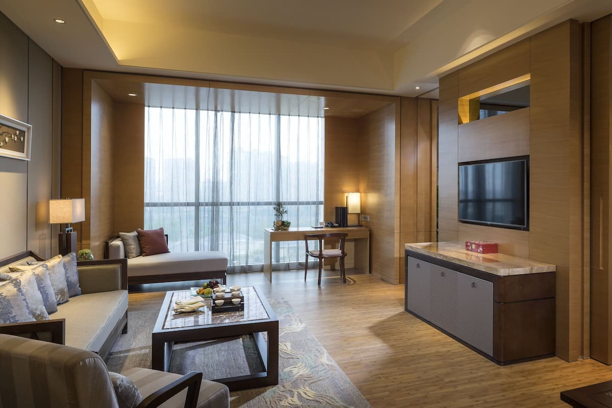 Habitació d'hotel moderna amb sofà, taula de dinar, zona de cuina, televisió i una finestra gran amb cortines.