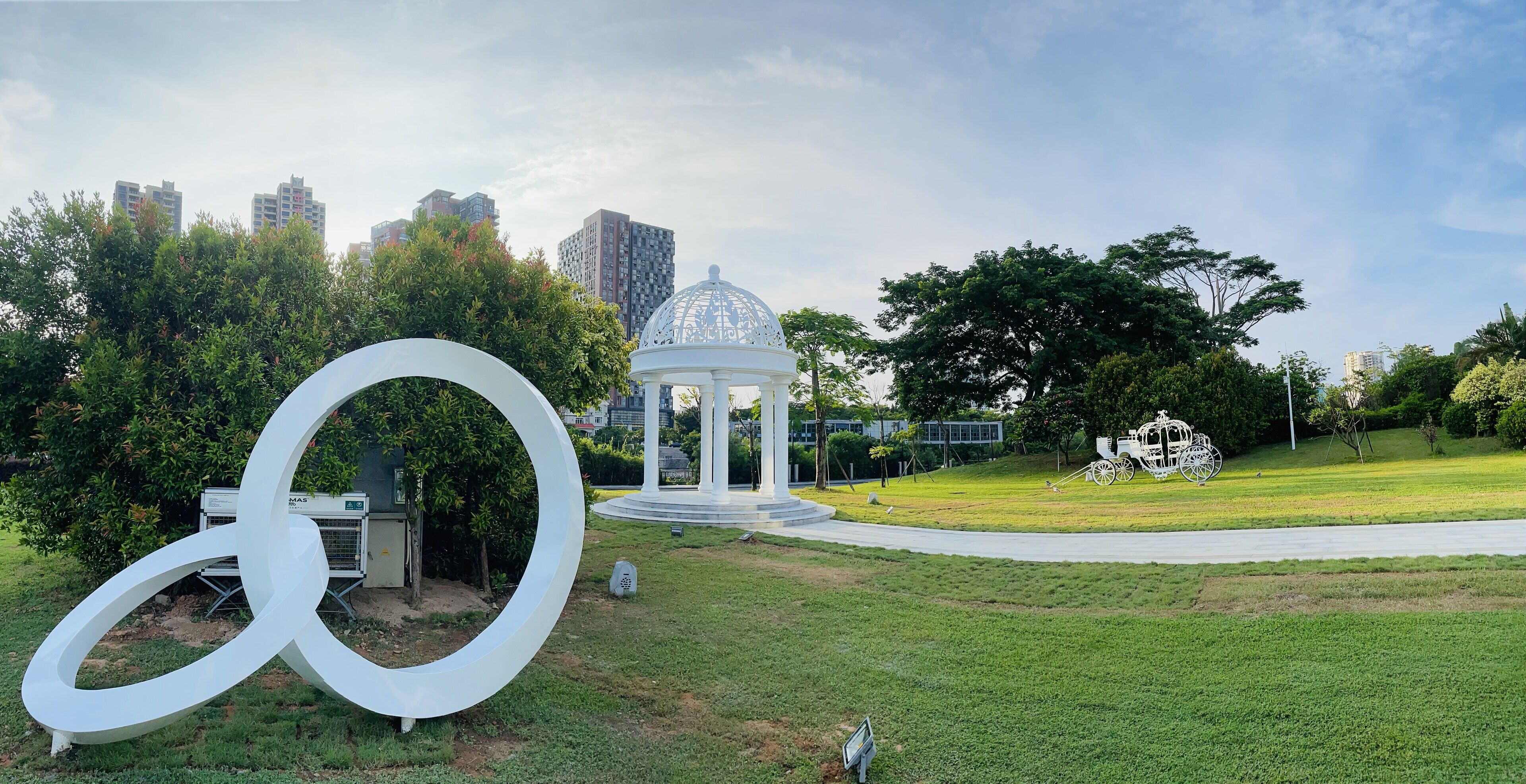 A park with a gazebo, a large white sculpture, and a cityscape in the background.