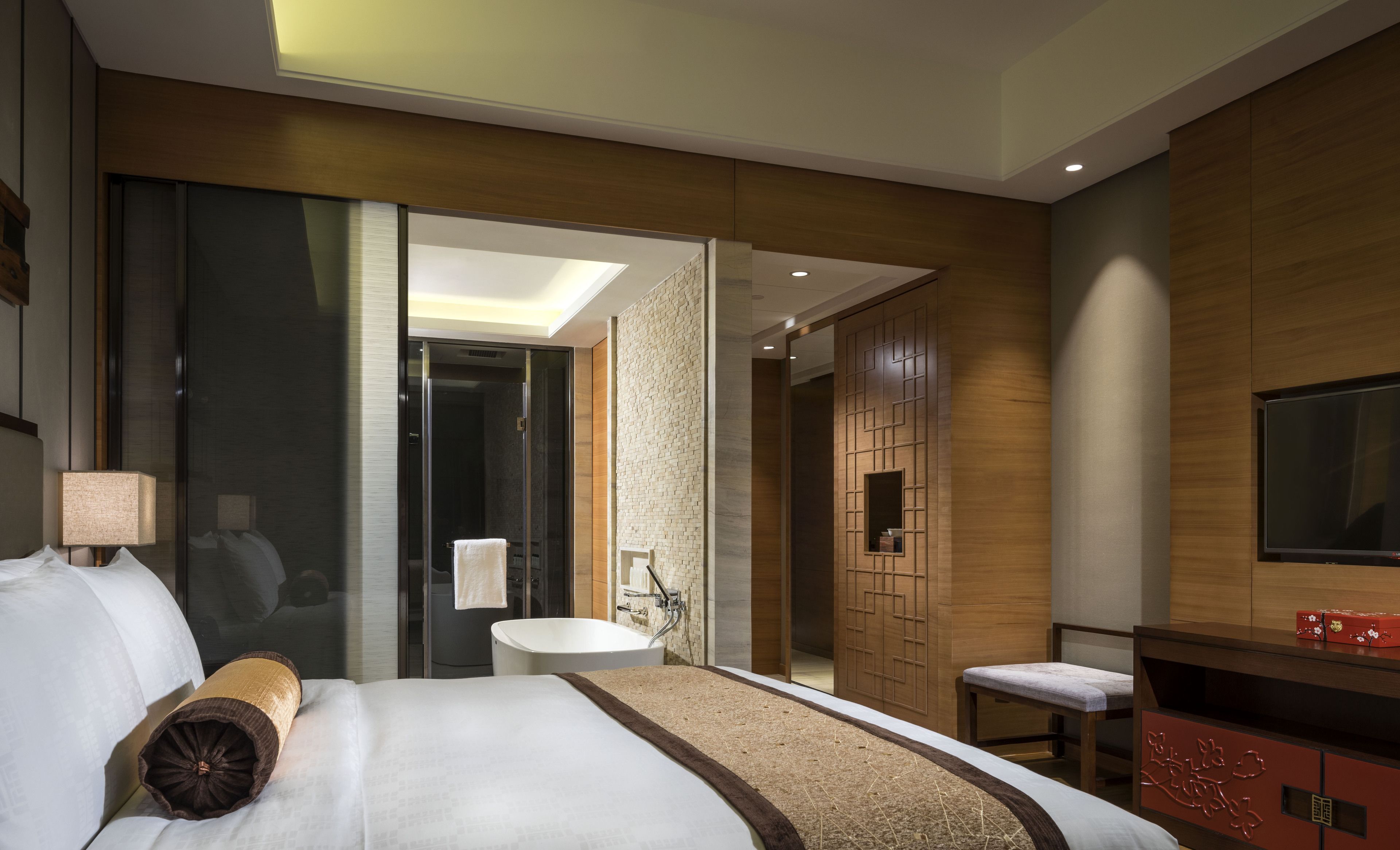 A modern hotel room with a large bed, a sitting area, a TV, and a bathroom with a bathtub and sink.