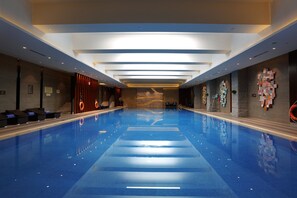 Ein großes Hallenbad mit klarem, blauem Wasser, umgeben von Liegestühlen und einem modernen Ambiente.