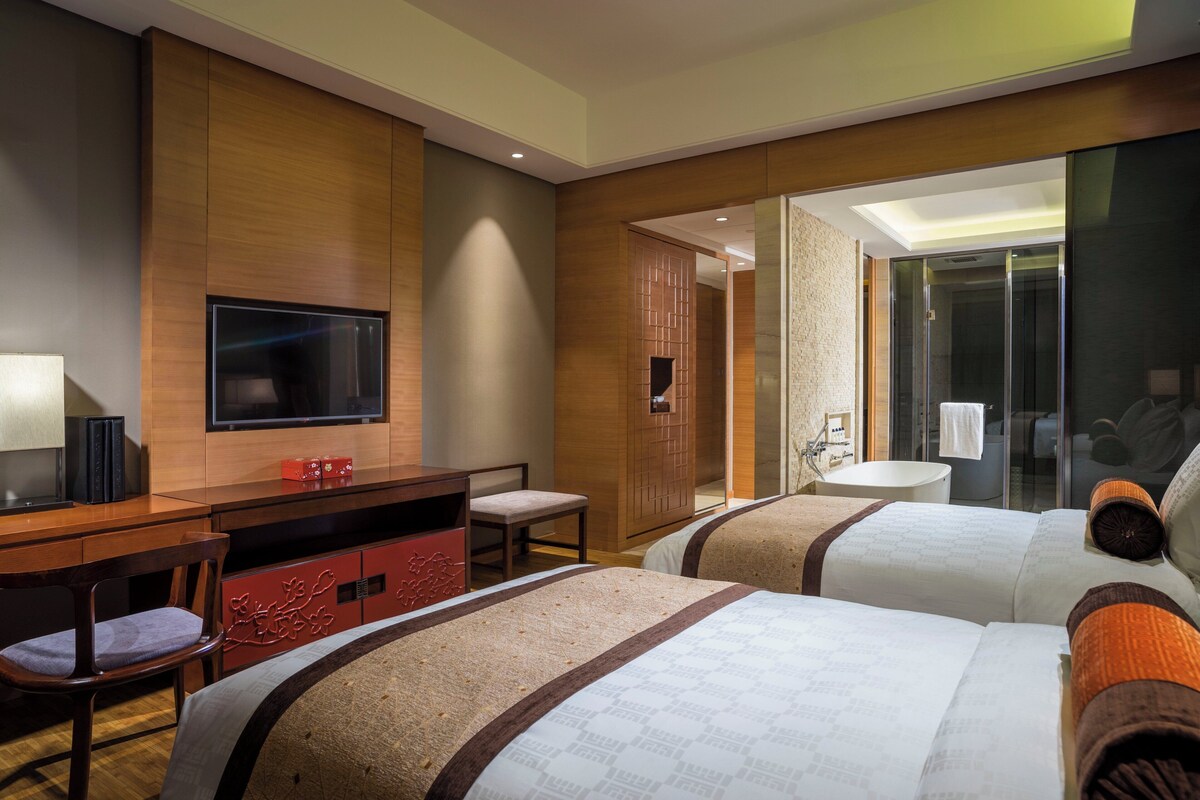 Ein Hotelzimmer mit Fernseher, Schreibtisch, Stuhl, zwei Betten und einem Badezimmer mit Dusche und Waschbecken.