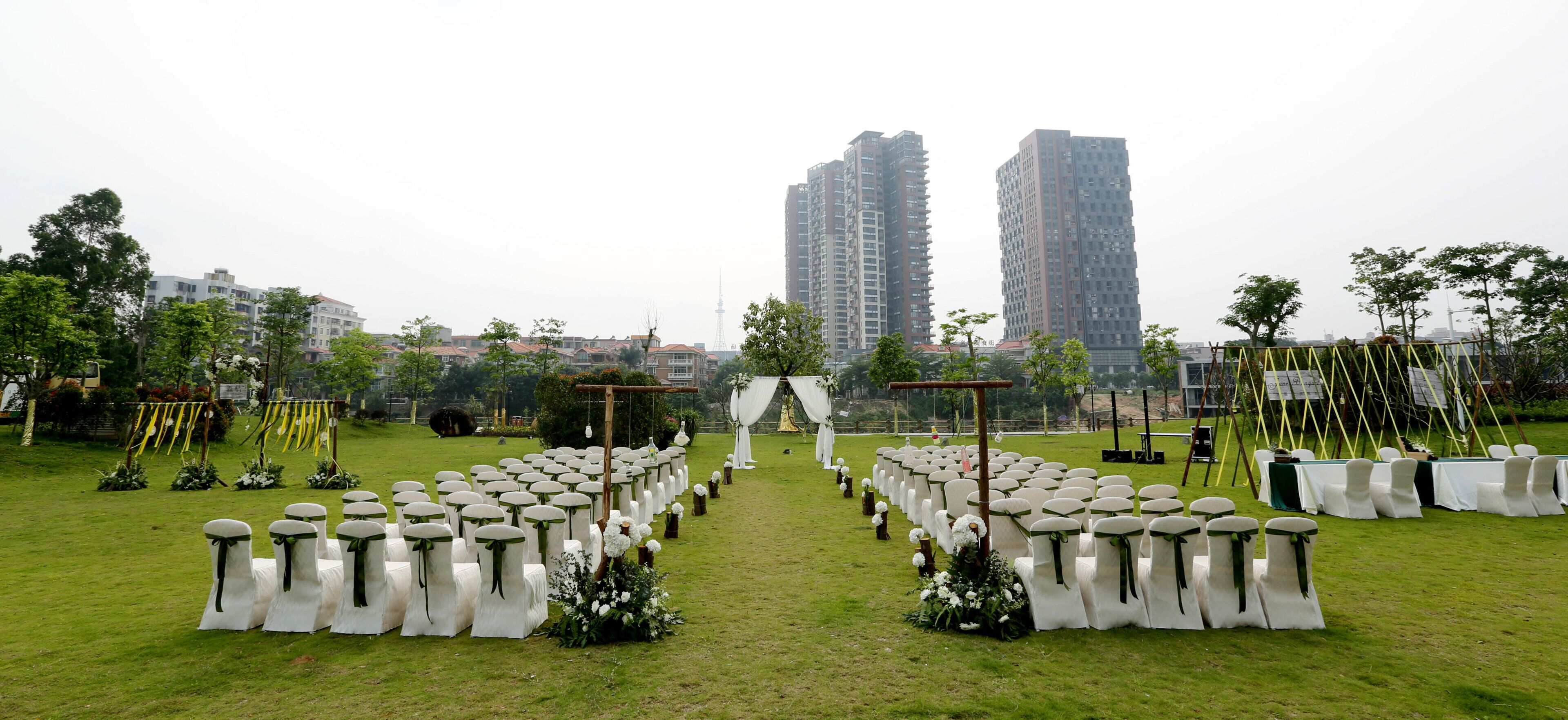 An outdoor venue with rows of white chairs and tables, a stage with a backdrop, and a view of tall residential buildings in the background.
