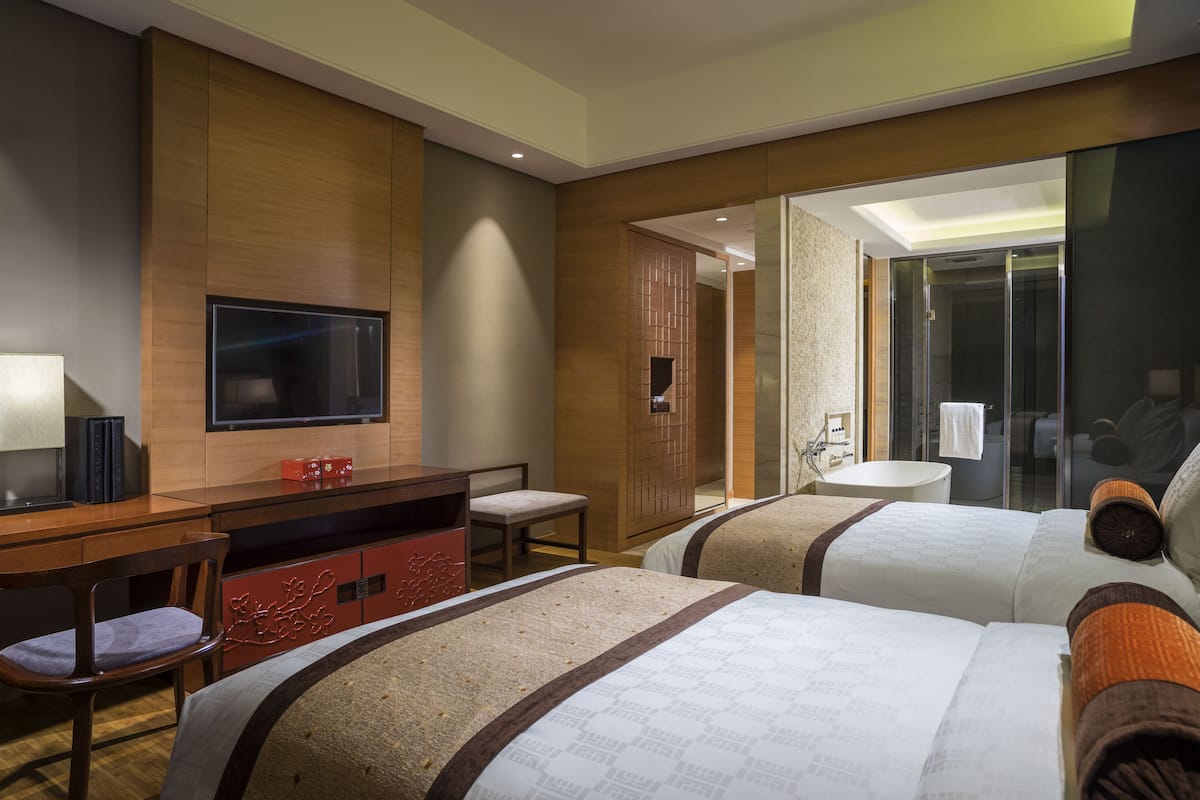 A hotel room with two beds, a TV, a desk, and a bathroom visible through a glass door.