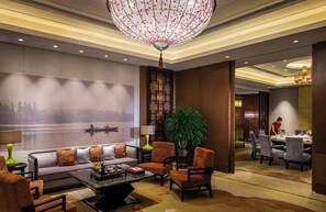 A modern hotel lobby with a large chandelier, a sofa set, a dining area, and a wall with a scenic painting.