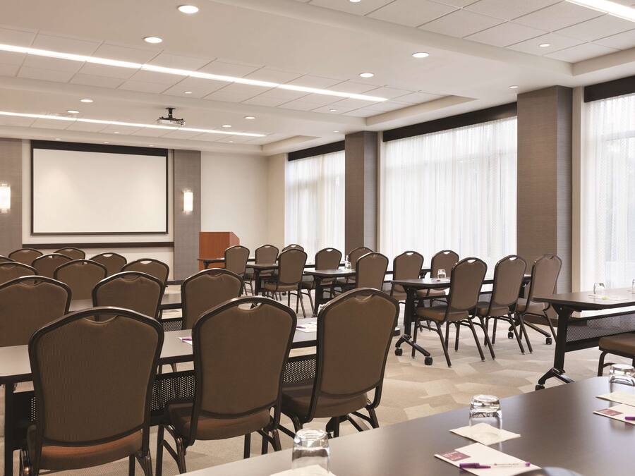 A conference room with multiple tables and chairs, a projector screen, and large windows with curtains.