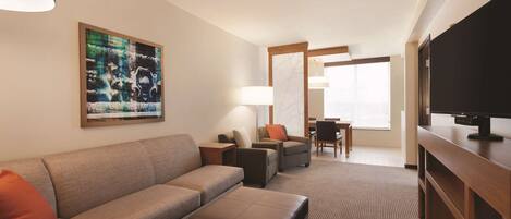 A modern hotel room with a sofa, ottoman, TV, and artwork on the wall.
