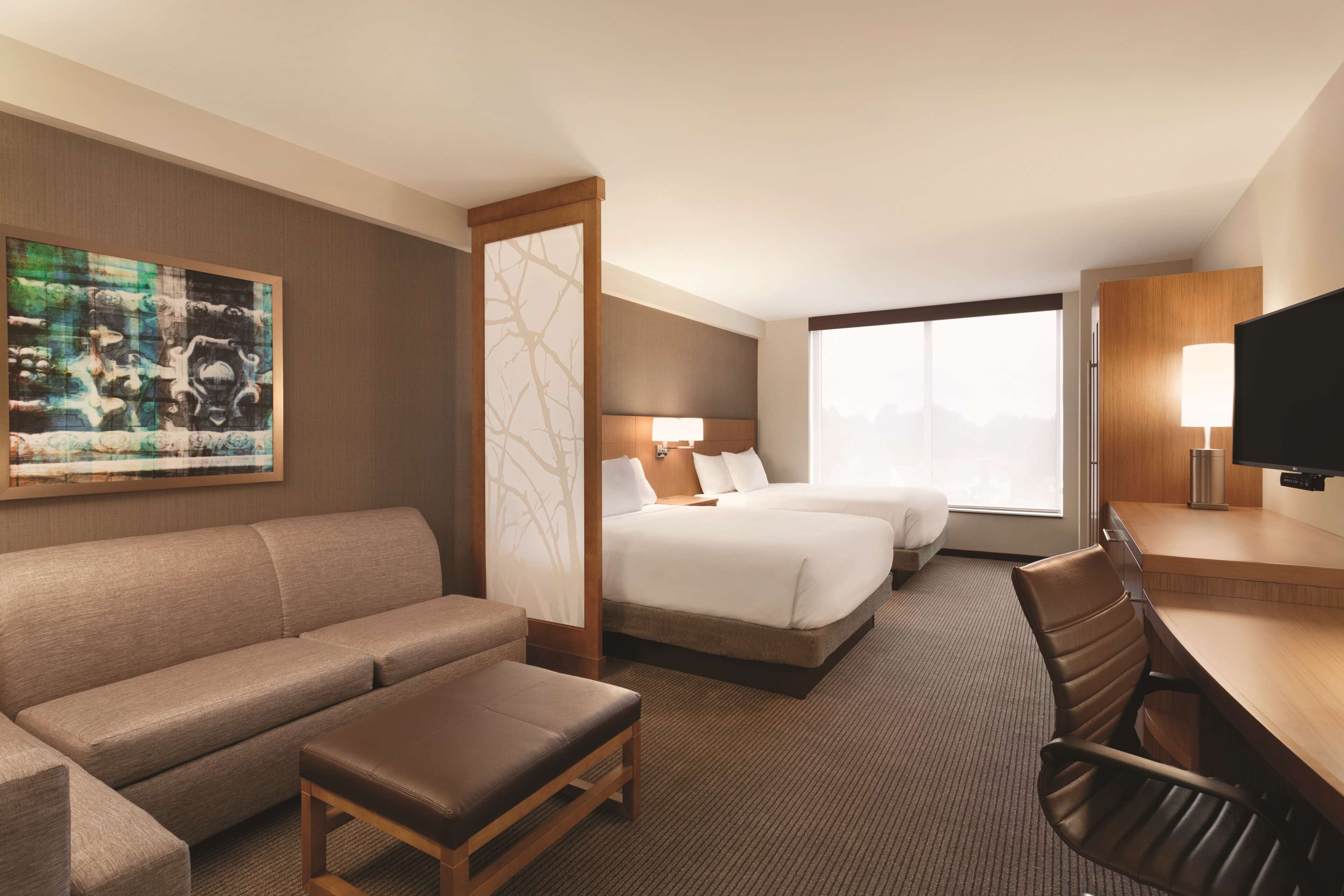 A hotel room with a sofa, two beds, a desk, a TV, and a painting on the wall.