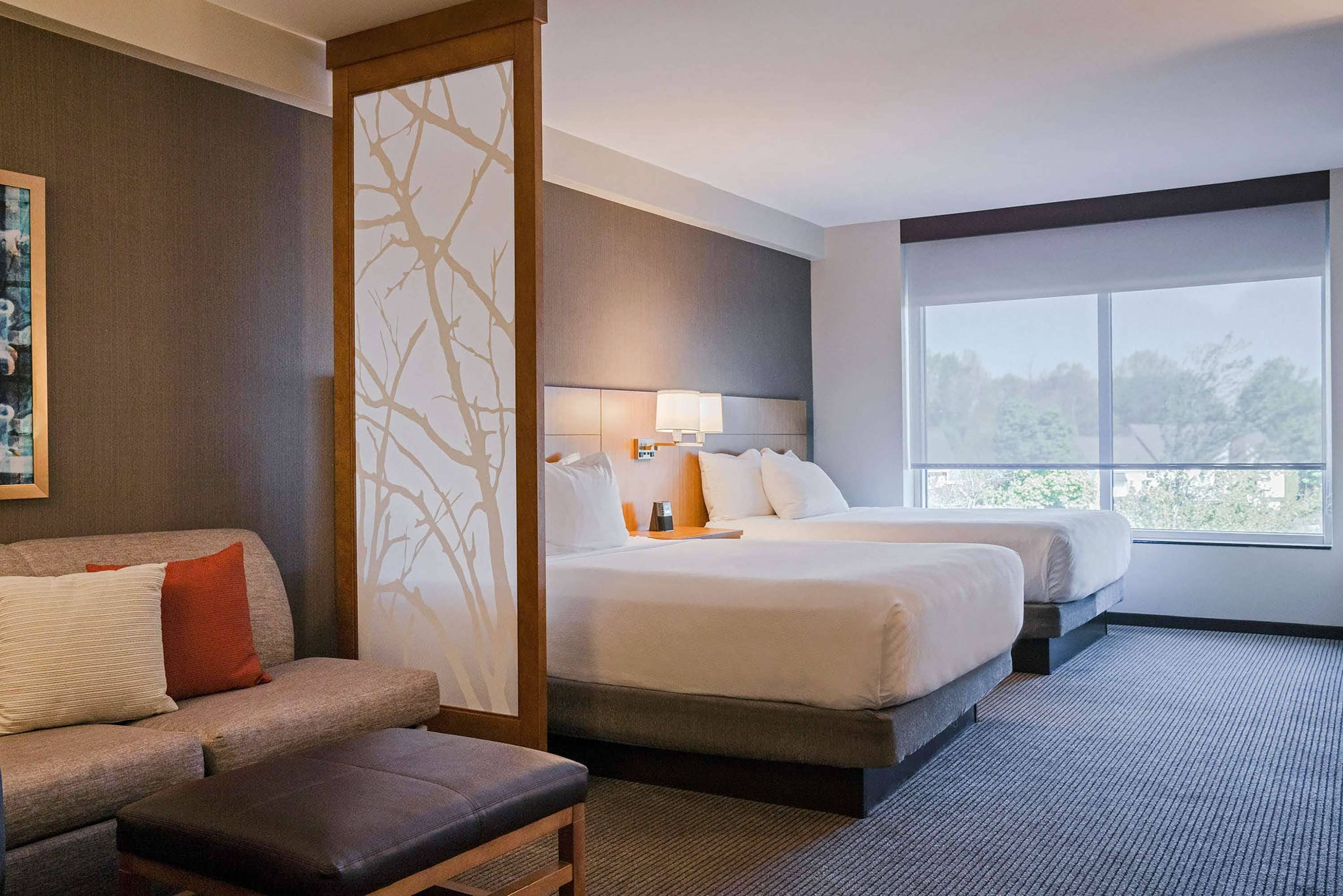 A hotel room with a large bed, a sofa, a small table, and a view of trees through the window.