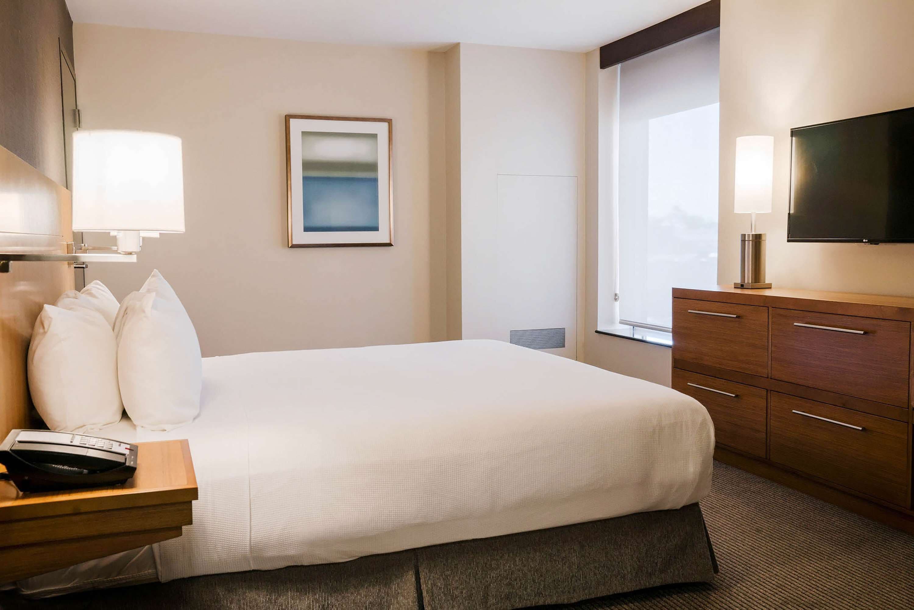 A hotel room with a large bed, a wooden nightstand with a telephone, a flat-screen TV mounted on the wall, and a window with a view.
