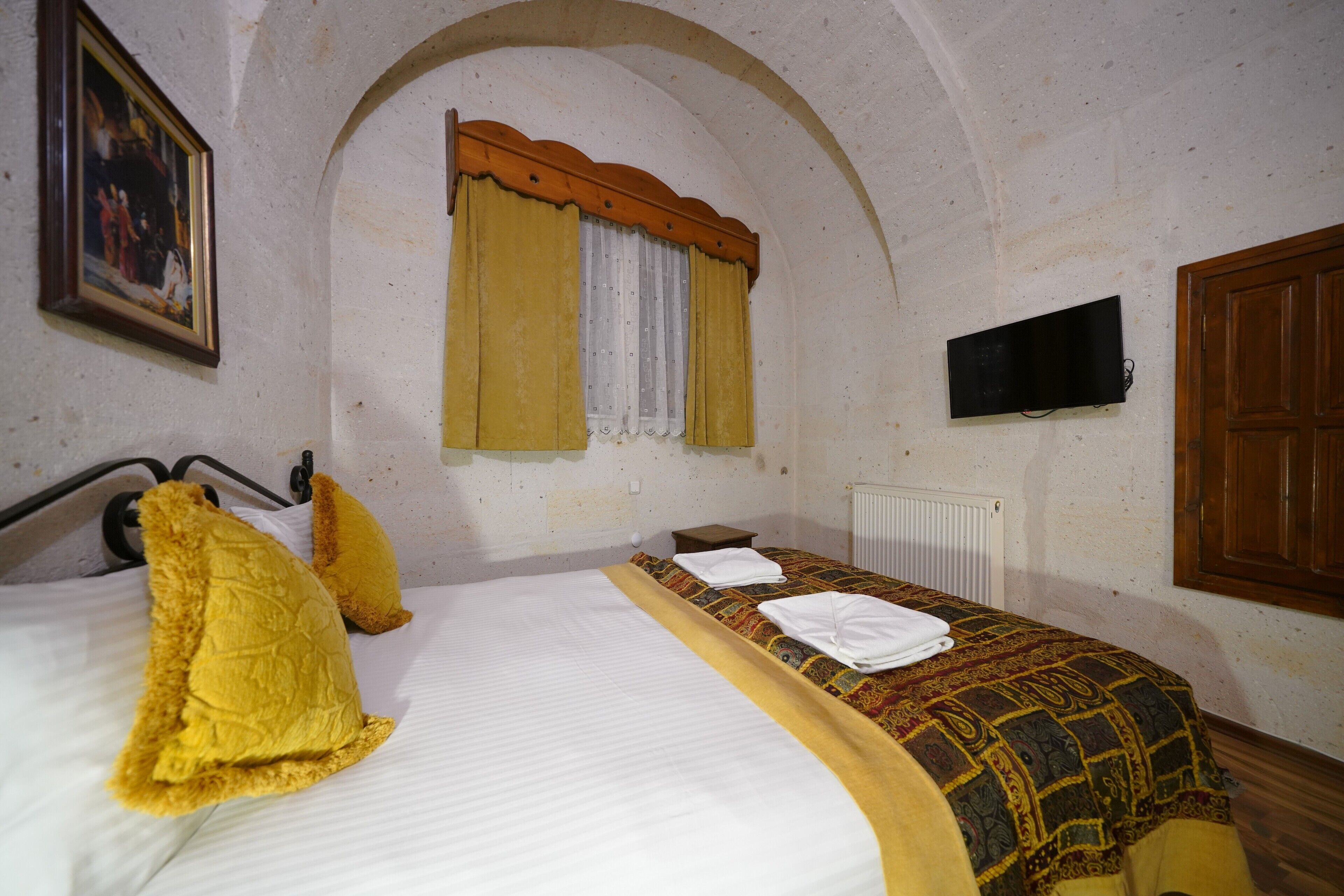 A bedroom with a bed, a window with curtains, a television mounted on the wall, and a wooden door.