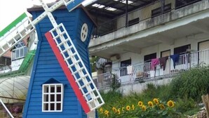 A blue windmill structure with a white door and windows, situated in a landscaped area with sunflowers and a building in the background. - Kea Valley Hotel Apartment (Brinchang)