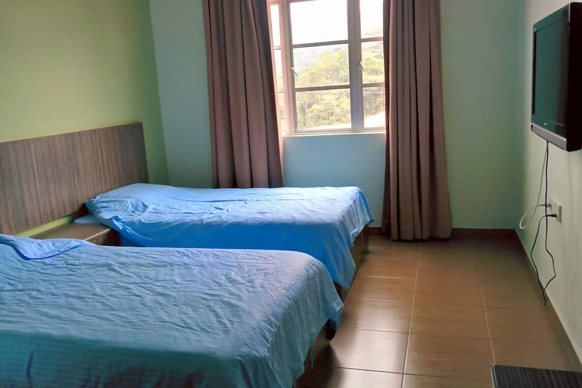Una habitación con dos camas individuales, una ventana con cortinas, un televisor de pantalla plana montado en la pared y suelo de baldosas.