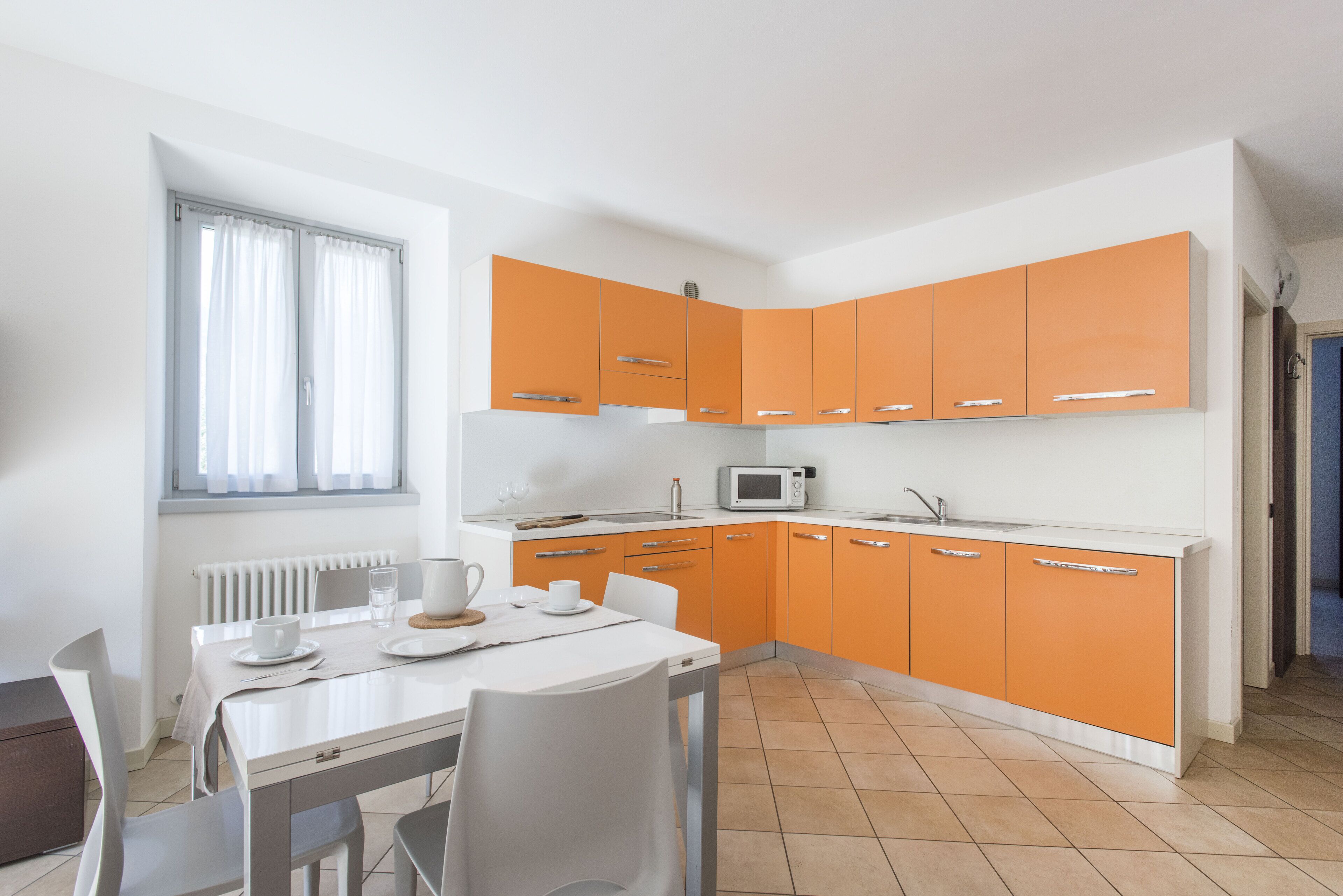 Una cocina moderna con armarios blancos y anaranjados, una mesa de comedor blanca y una ventana con cortinas blancas.