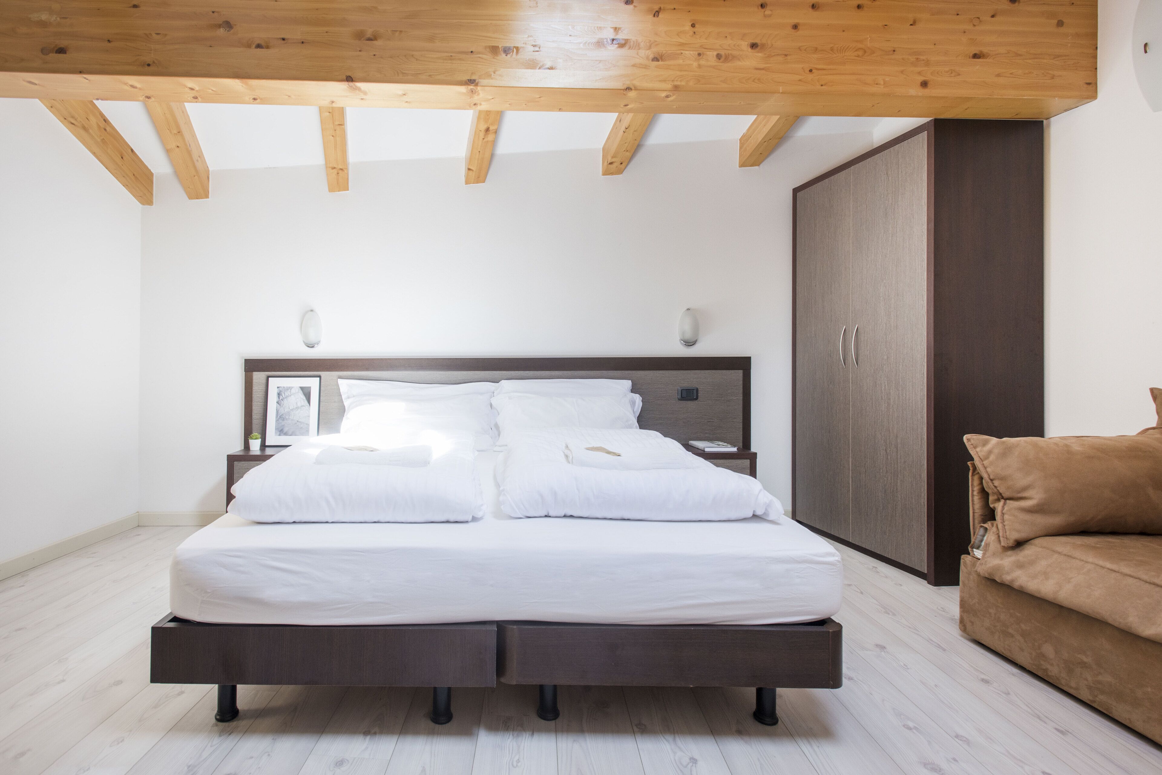 Un dormitorio con techo de madera, una cama con ropa de cama blanca, una mesita de noche, un armario y un sofá marrón.