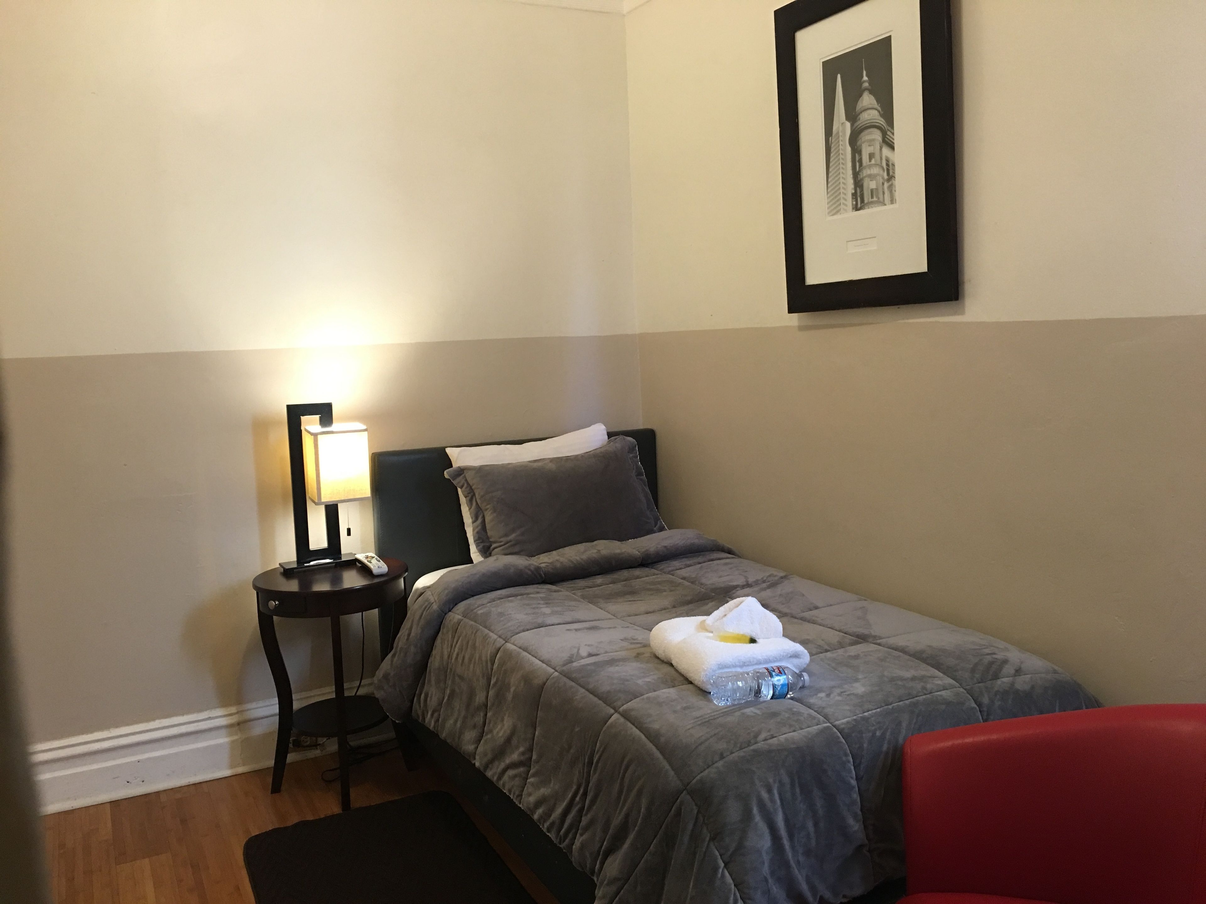 A neatly made bed with a grey duvet, a small bedside table with a lamp, a framed picture on the wall, and a red chair.