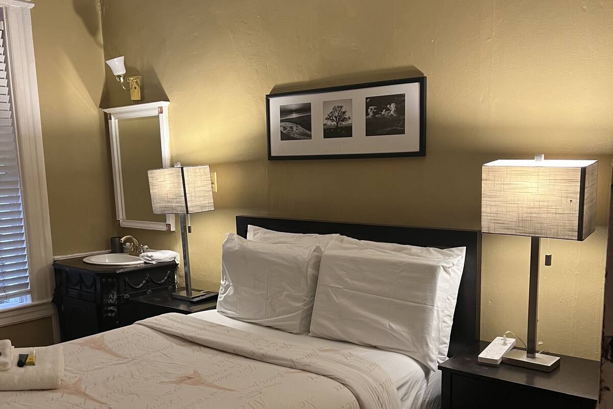 A hotel room with a bed, bedside tables, a nightstand, a mirror, and a wall-mounted picture frame.