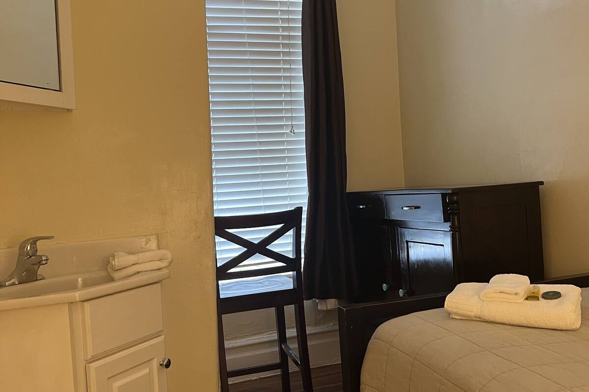A hotel room with a bed, a desk, a chair, a window with blinds, a sink, and a mirror.