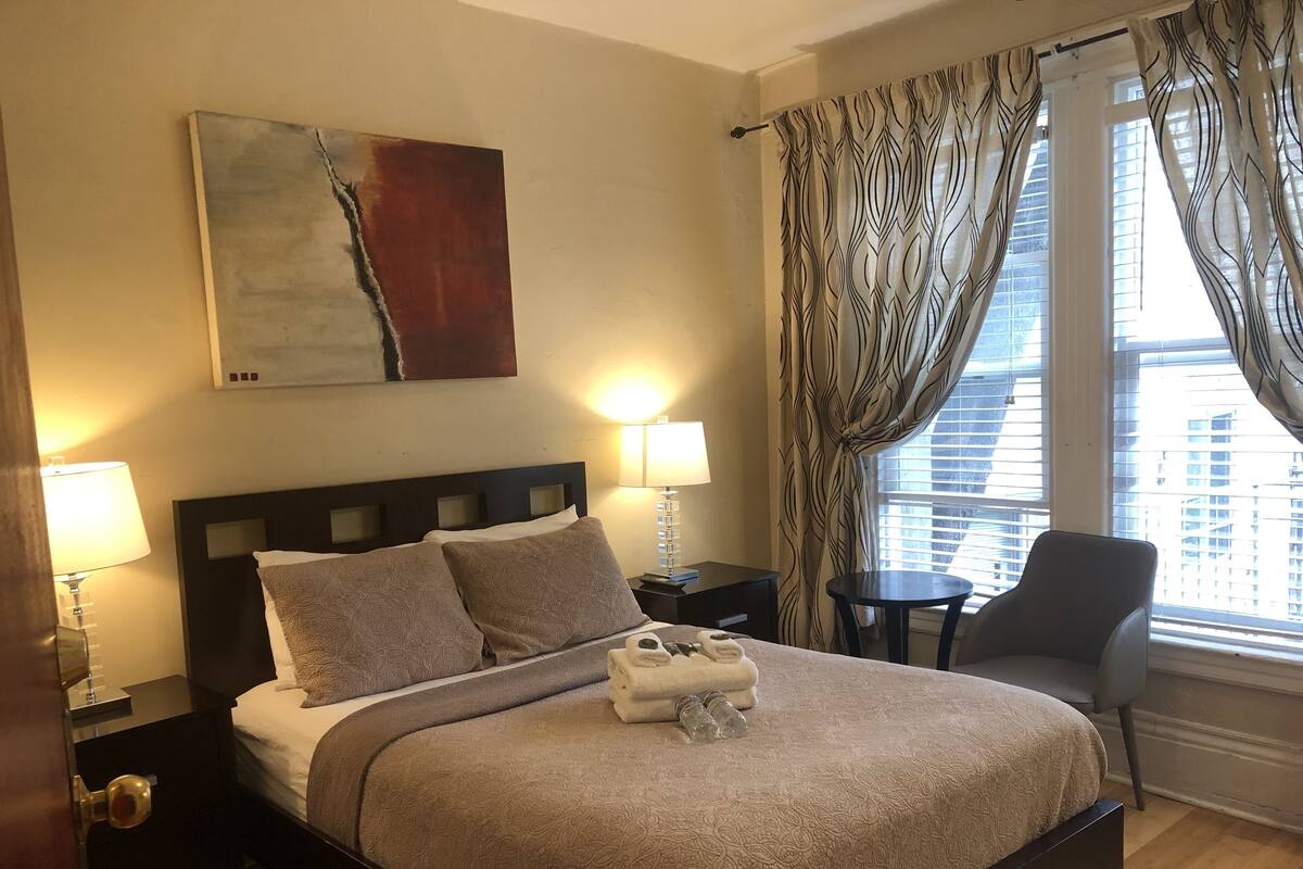 A hotel room with a large bed, two bedside tables, a chair, a painting on the wall, and a window with curtains.