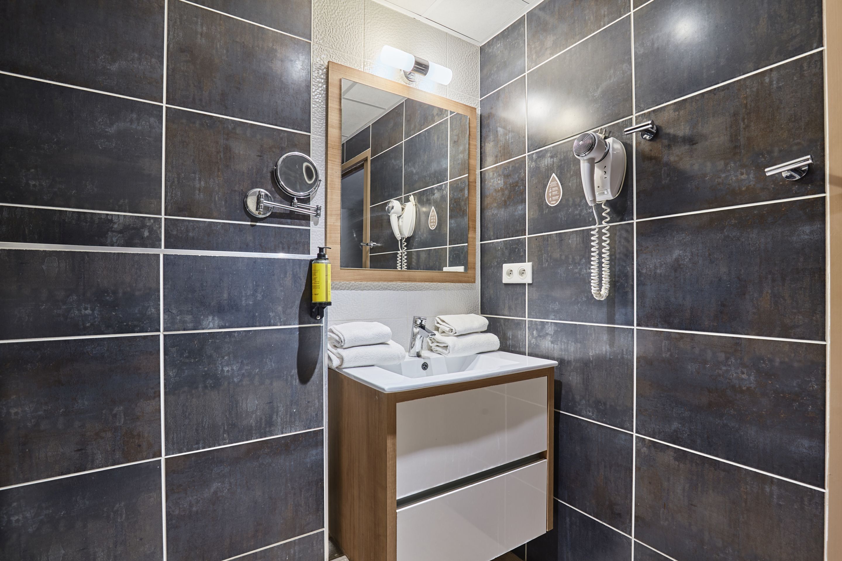 A bathroom with dark tiled walls, a wooden vanity, a mirror, a hairdryer, and a towel dispenser.