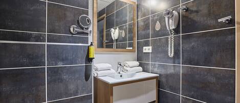 A bathroom with dark tiled walls, a wooden vanity, a mirror, a hairdryer, and a towel dispenser.