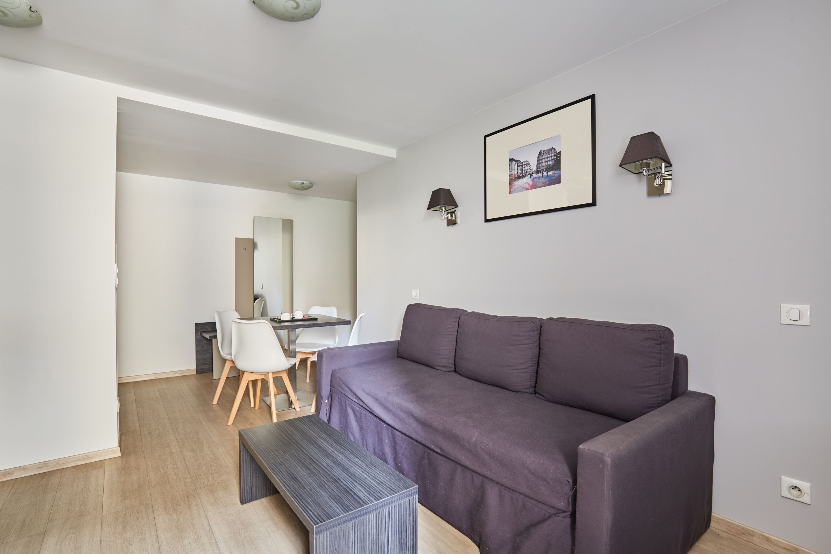 A modern living room with a dark-colored sofa, a small wooden bench, and a dining area with a table and chairs.