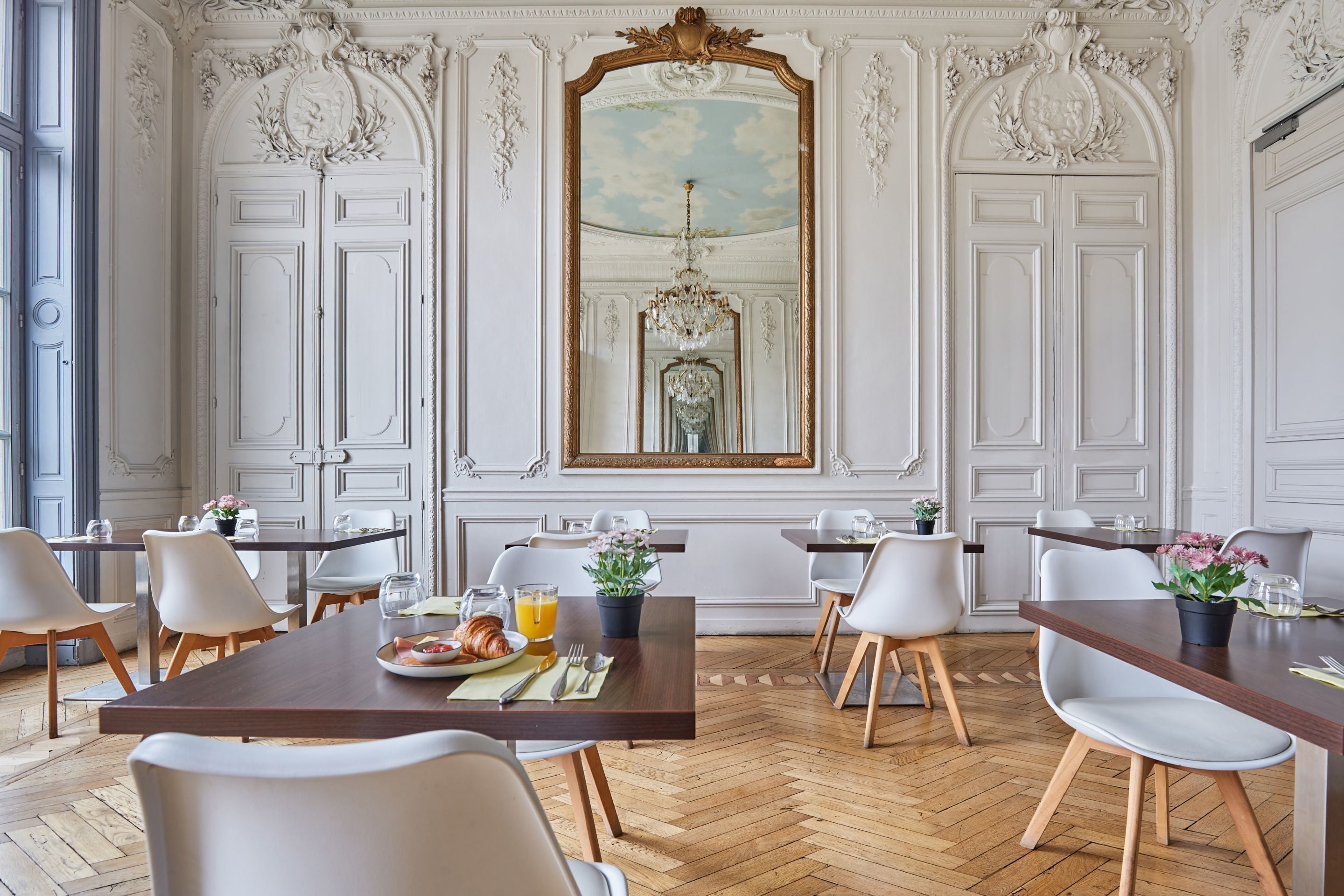 Comedor con piso de madera en patrón de espiga, sillas blancas, una mesa central con alimentos para el desayuno y un espejo grande y decorativo sobre la puerta.