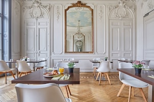 Uma área de refeições com piso de madeira em padrão herringbone, cadeiras brancas, uma mesa central com itens de café da manhã e um espelho grande e ornamentado sobre a porta.