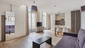 Un salon moderne avec un canapé, une table basse et un téléviseur. On aperçoit un coin repas avec des chaises et, au fond, une cuisine.