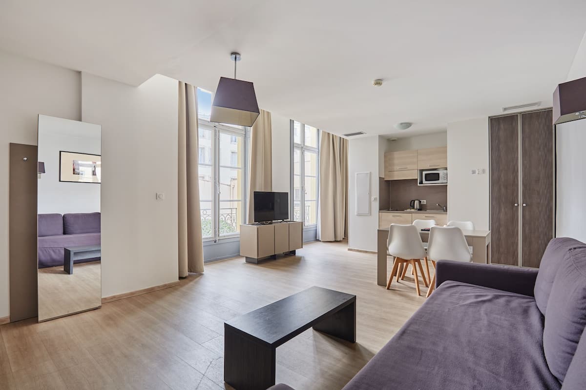 Ein modernes Wohnzimmer mit einer Couch, einem Couchtisch und einem Fernseher. Im Hintergrund befindet sich ein Essbereich mit Stühlen und eine Küche.