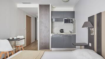 Ein kompaktes Hotelzimmer mit Bett, Schreibtisch, Stuhl, einer Küchenzeile und einer Tür zu einem weiteren Raum.