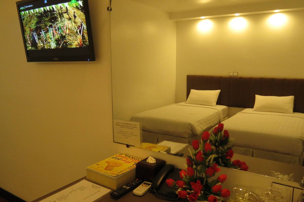 Ein Hotelzimmer mit zwei Betten, einem an der Wand befestigten Fernseher, einem Schreibtisch mit einem Buch, Fernbedienungen, einer Vase mit roten Blumen und einem Spiegel, der das Zimmer reflektiert.