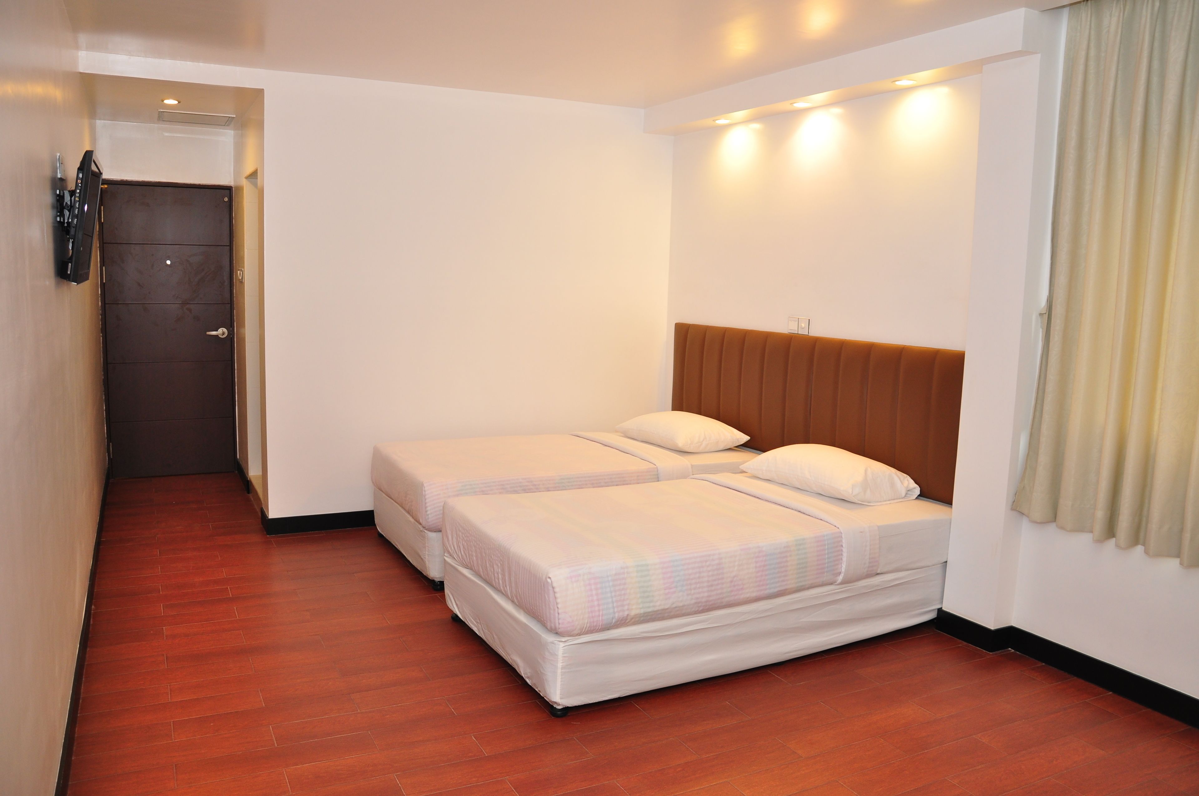 A hotel room with two beds, wooden flooring, a brown headboard, and a door with a security device.
