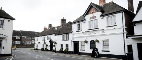 A row of white buildings with black trim, including a pub named "The Crown Inn" with hanging flower baskets and a blackboard sign.
