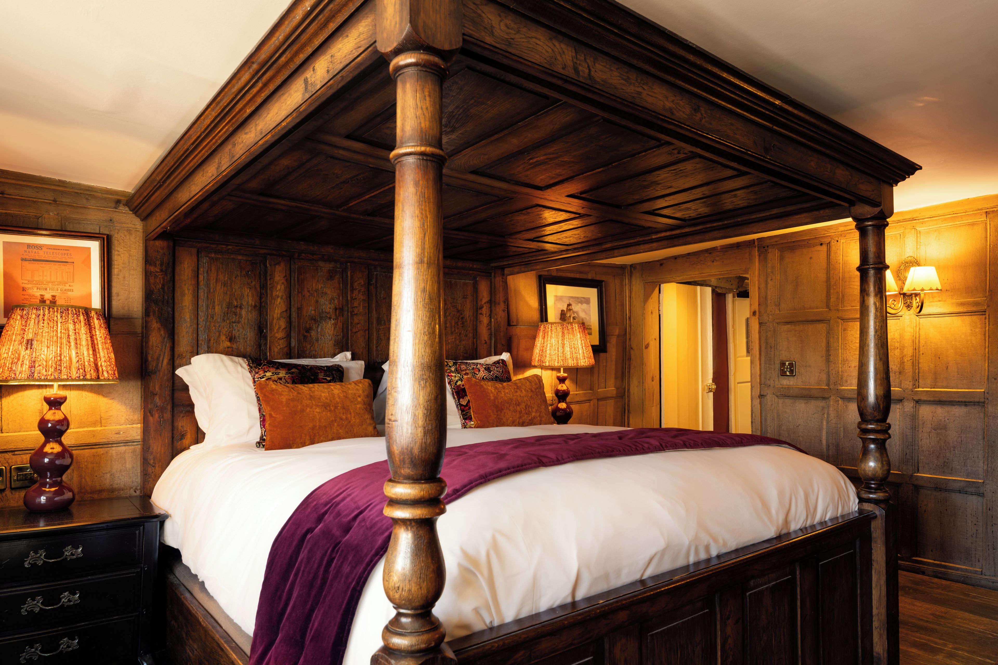 A traditional four-poster bed with a wooden headboard, a white bedspread, and a purple throw blanket. There are two bedside lamps with lampshades, a framed picture on the wall, and a wooden dresser with drawers.