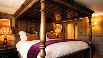 A traditional four-poster bed with a wooden headboard, a white bedspread, and a purple throw blanket. There are two bedside lamps with lampshades, a framed picture on the wall, and a wooden dresser with drawers.